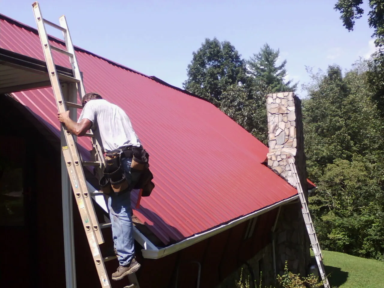 Roofer on a ladder beside a red metal roof and stone chimney — Soffit & Fascia Repair by Roofing Brothers in Newburgh